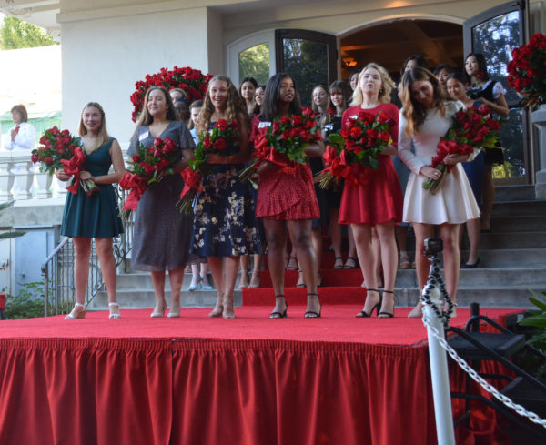 Mia Thorsen stands on red-carpeted dais with five other members of the Royal Court, holding red roses