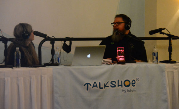 DeltaMaker Marketing and Outreach Manager Andy Stetzinger talks to host Dot Cannon at table with mics and "Talkshoe" on a white tablecloth, beside a lighted "Over Coffee" luciet sign