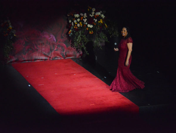 Lynette Romero in flowing red gown, steps onto red carpet onstage with a mic in her hand for Rose Queen® coronation