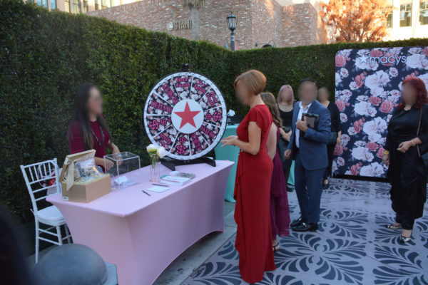 Woman in a red dress smiles as she stands near outdoor table with Macy's logo and prize wheel in background, with woman behind the table and other attendees in evening clothes in the background