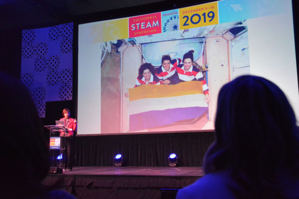 Ellen Ochoa stands onstage next to slide of herself with two fellow astronauts in training during California STEAM Symposium