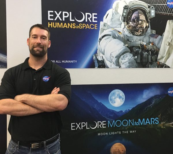 Matt Wittal stands near a NASA poster with "Explore Moon to Mars" and a picture of an astronaut in a spacesuit