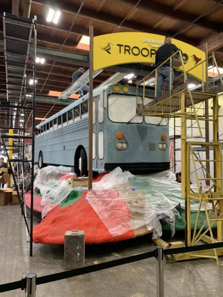 Volunteer stnds on scaffolding to glue materials on a yellow "Troop Zero" banner above a bus