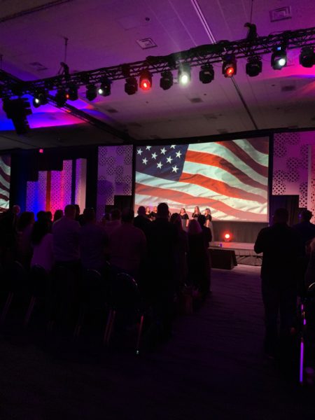 Audience stands as Bella Voce sing National Anthem in front of a waving American flag projected on video