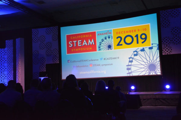 Audience in darkened auditorium looks at slide advertising "California STEAM Symposium 2019"