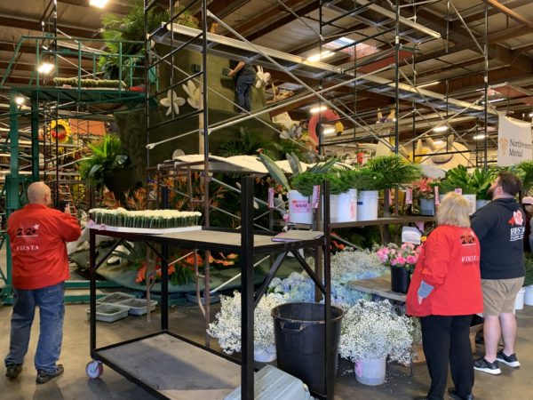 Fiesta Parade Floats staffers in red jackets stand near float, preparing to decorate it with green branches from nearby containers