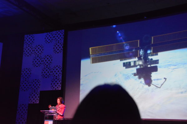 Dr. Ellen Ochoa speaks from lectern of California STEAM Symposium with slide of ISS as seen from space shuttle in the background