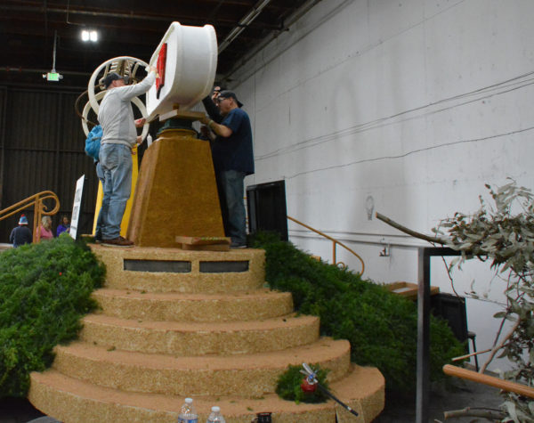 Four volunteers glue something on either side of a white sign at the top of six steps on the football float, where the steps are partially covered with greenery