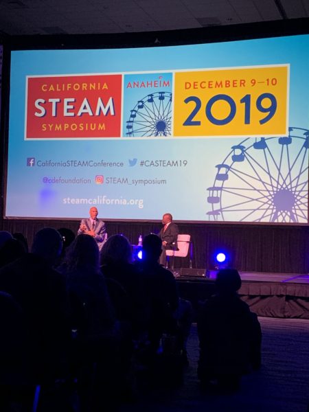 Long shot of Superintendent Tony Thurmond interviewing Kareem Abdul-Jabbar in front of "California STEAM Symposium 2019" slide