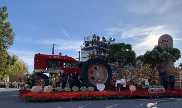 Chipotle float features a huge red tractor with "Chipotle" on it, surrounded by barnyard animals made of plant materials, as it passes the reviewing stand