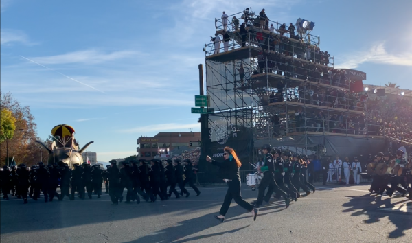 Band coordinator Emiko Christensen waves as Green Band members take the corner at a run near the Rose Parade reviewing stand