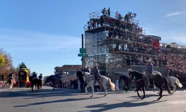 Riders in Mexican cowboy outfits wave from the backs of a high-stepping white horse and black horse