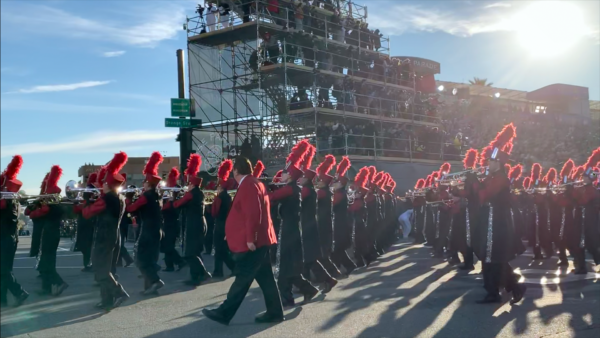 Rancho Verde band, in red and black uniforms, playing trombones and trumpets, walk by the reviewing stand with their band director