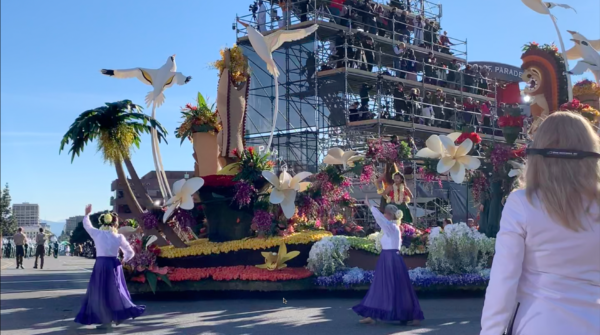Outwalkers in white blouses and long skirts walk near Sierra Madre float depicting a Hawaiian landscape with tikis, volcanoes and outsized plumeria flowers