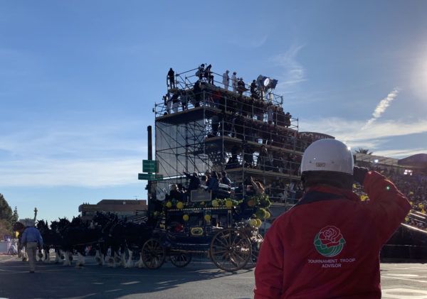 Red-jacketed Tournament of Roses supervisor salutes as Pasadena Mayor Terry Tornek and his family wave from the top of replica of an 1880s coach pulled by a team of black-and-white Percherons