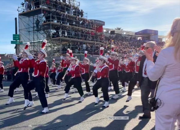 Wisconsin bandmembers perform a high step in their red-and-black uniforms with white spats while playing trumpets