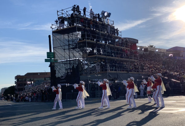 Seven PCC Herald Trumpeters, in white slacks and capes with red tops, blow elongated trumpets next to the review stand