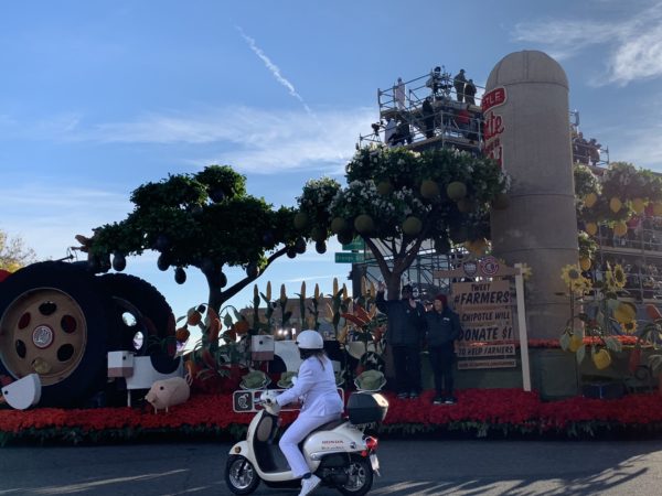 Sign on back of Chipotle float says, "Tweet "farmers" and we will donate $1 to support farmers" as two farmers wave from the float