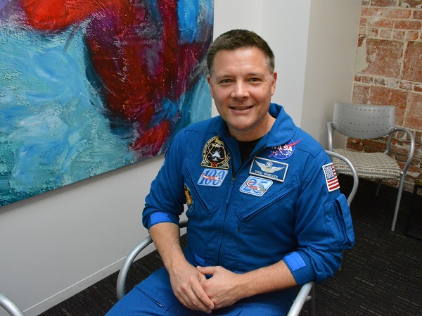 NASA Astronaut Doug Wheelock, in his blue astonaut flight suit, smiles from an office chair in Pasadena's Cross Campus facility.
