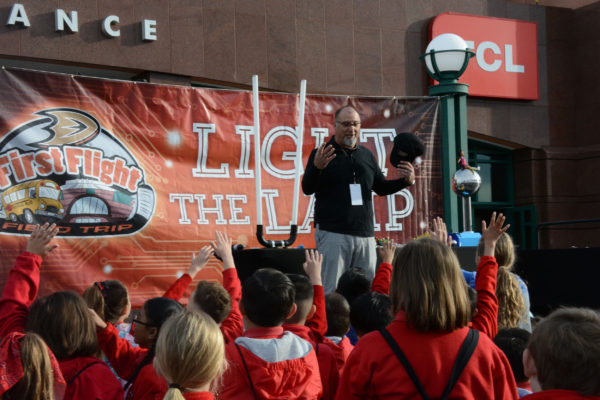 Elementary students around outdoor stage raise hands to volunteer as Paul speaks in front of "Light the Lamp" banner