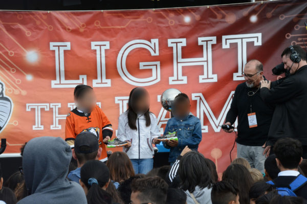 Paul talks three elementary students through lighting up shoes onstage as cameraman captures the moment