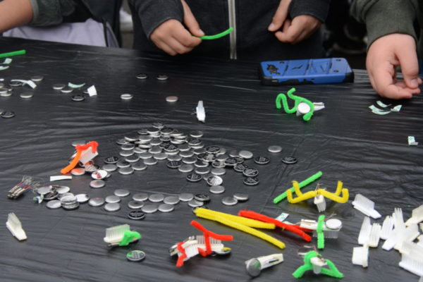 Toothbrush "bugs" on table, surrounded by small hands and piles of components, during "First Flight Field Trip"