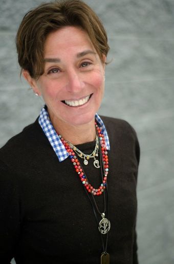 Living Popups CEO Cheryl Bayer, in a black sweater and with a red beded necklace and silver chain around her neck, smiles against a gray background
