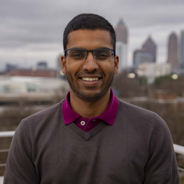 Aditya Vishwanath, in. ared shirt and brown sweater, smiles against a blurred city skyline background