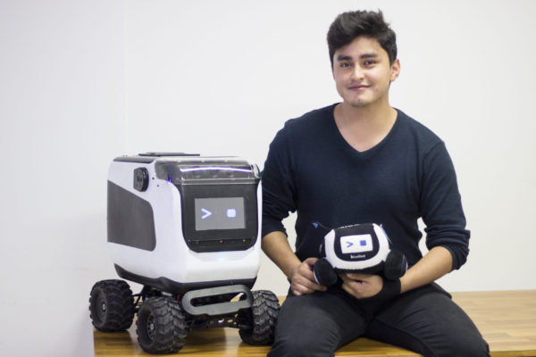 Alejandro Otalora kneels on the floor next to Kiwibot four-wheeled robot as he holds another, smaller robot model.