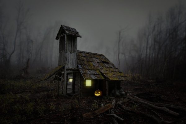 Creepy wooden house with tall cupola stands alone in a field with lights on in the windows and a lighted Jack-o-Llantern near the door