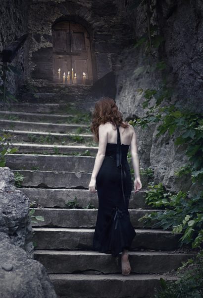 A red-haired woman in a long black gown climbs ancient stone steps to a closed wooden door with a cross symbol on it.