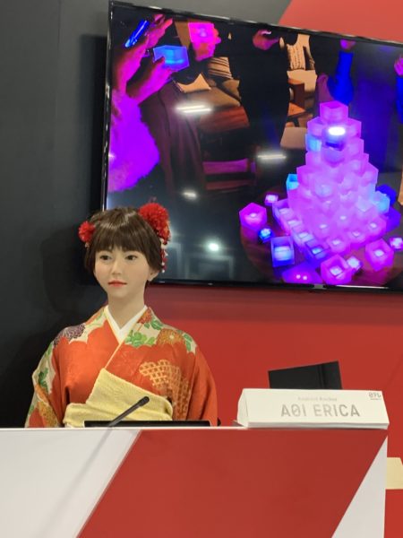 Humanoid robot "Erica', dressed in a kimono, stands behind a counter in CES 2019's Eureka Park startup area.