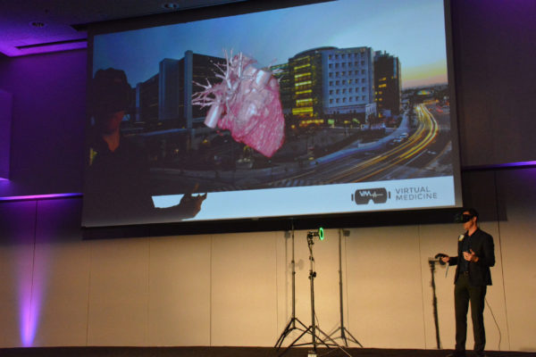 Dr Brennan Spiegel onstage in VR headset with a screen showing overlay of heart over Cedars-Sinai building