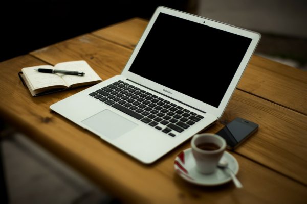 Laptop computer sits on wooden desk near cup of coffee, phone and small notebook with a pencil resting on it