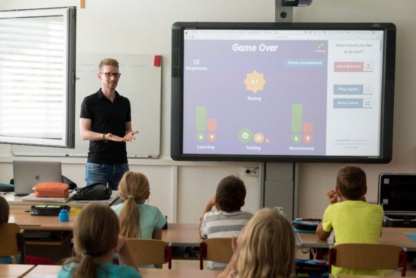 Young male teacher stands before a classroom of approximately first-grade students with a screen showing "Game Over"