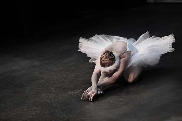 Ballet dancer, in a white tutu, strikes a ballet pose, kneeling with her arms crossed in front of her.