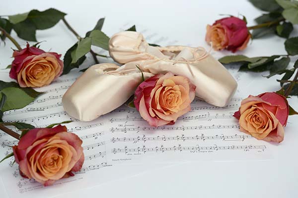 White satn ballet shoes crossed atop an orange rose on sheet music, surrounded by four other orange roses