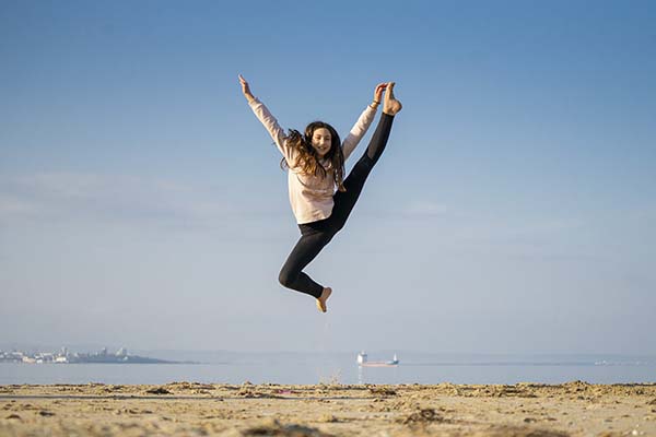 Young girl leaps into the air on a beach with background of ocean, sunny ay and island behind her