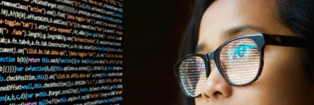 An Asian woman programmer with brown hornrimmed glasses looks at a screen covered with lines of code