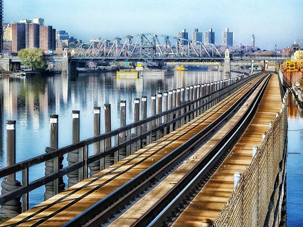 Skyline of the Bronx from wooden subway track on trestle over a blue body of water with extension bridge in the distance.