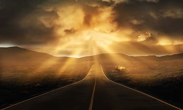 Sunrise rays break through dark clouds as a road leads forward with a mountain in the distance.