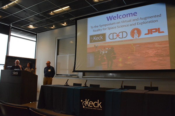 three symposium organizers at a lectern with "wecome" slide on a screen