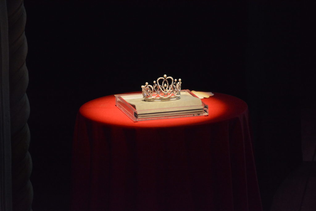 Crown for 101st Rose Queen sits on a white suchion atop a spotlighted red table onstage