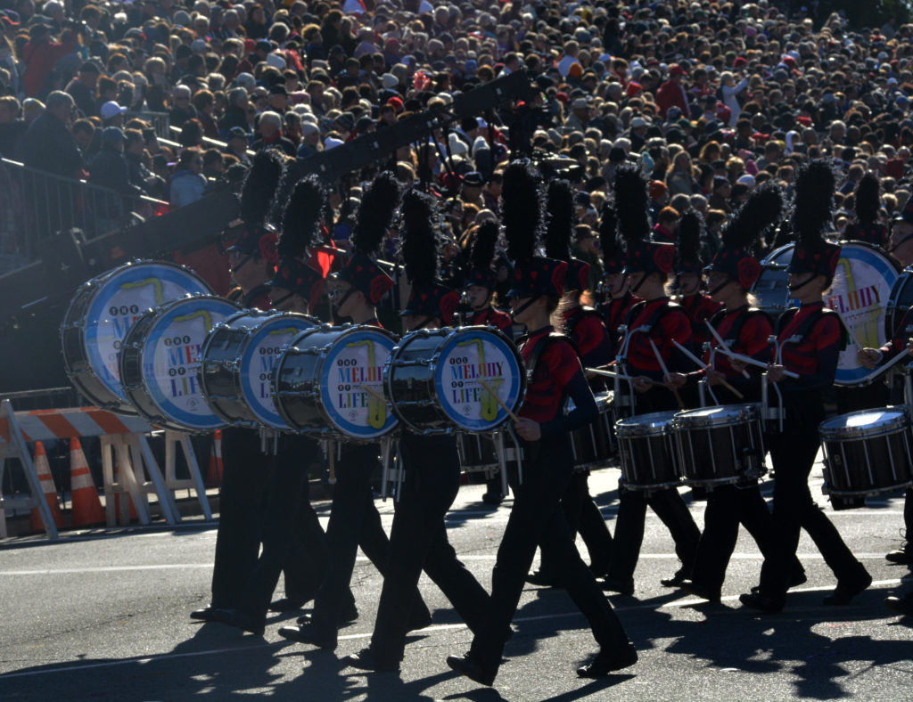 Band in red jackets with black trousers and black plumes on their hats marches past audience with drums reading "The Melody of Life"