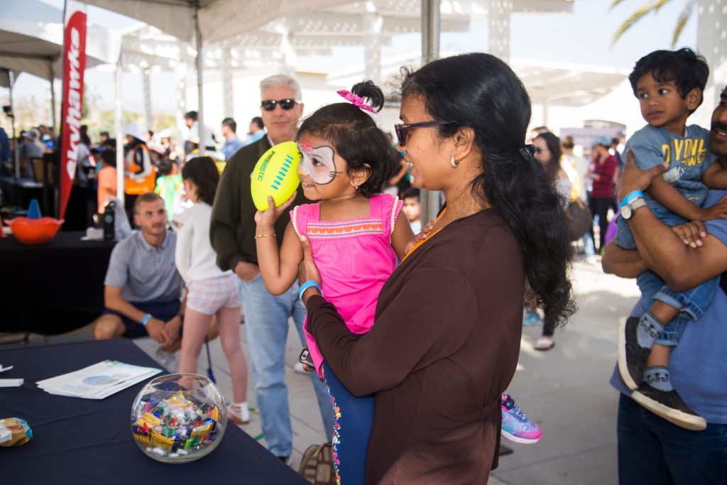 Small girl with face painted prepares to toss a yellow rubber football as her mother holds her inside OC STEAM Fest building