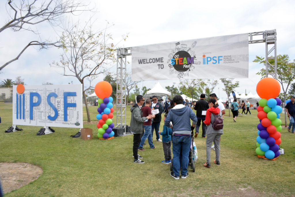 Adults and children enter park area under a white banner that says, "Welcome to STEAM Fest", next to a sign that says "IPSF"