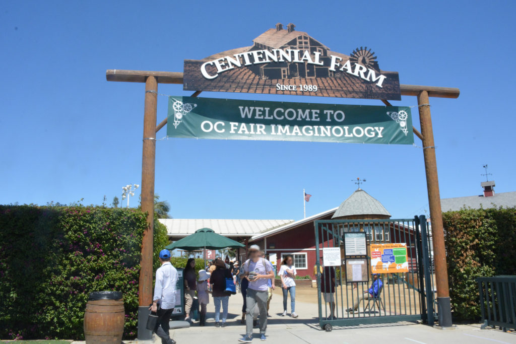 Attendees enter OC Fairgrounds under a wooden-supported sign that says "Centennial Farm", which is decorated with a green "Welcome to Imaginology" banner