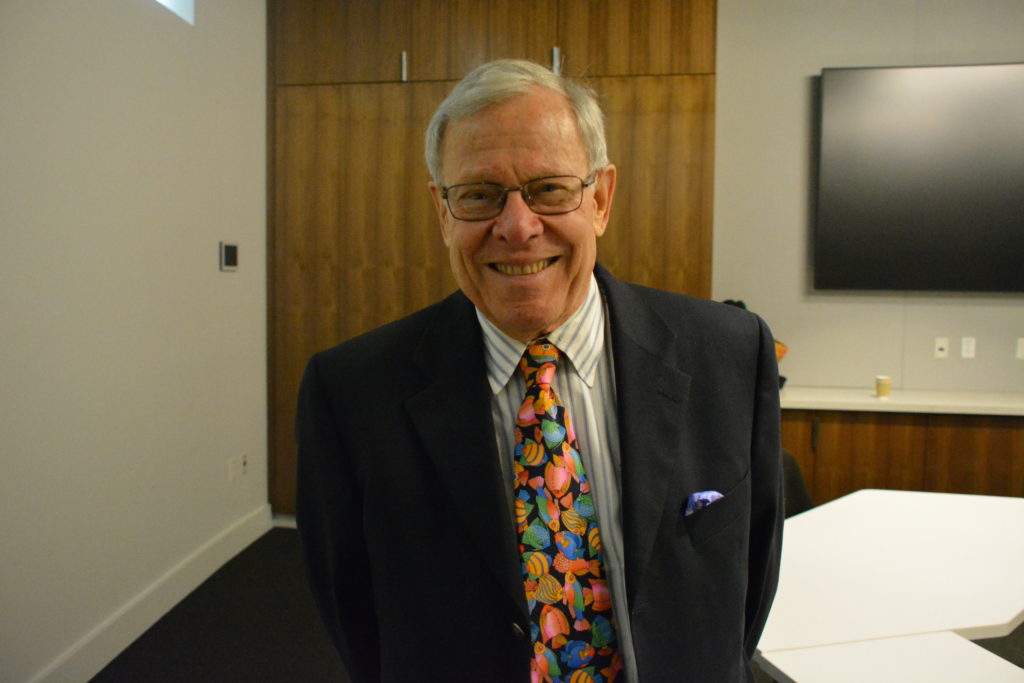 Dr. Jerry Schubel, President and CEO of Aquarium of the Pacific, smiles as he stands in a conference room wearing a black suit and a tie covered with brightly-colored tropical fish.
