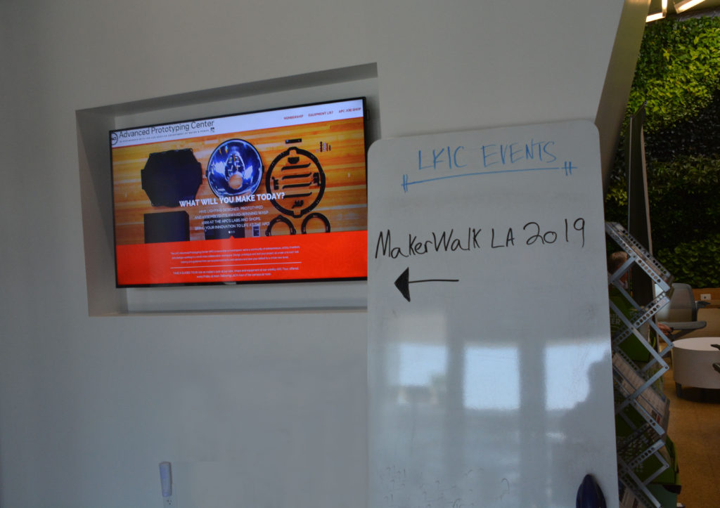 Whiteboard with "MakerWalk 2019" and arrow on in next to video screen projecting, "What will you make today?"