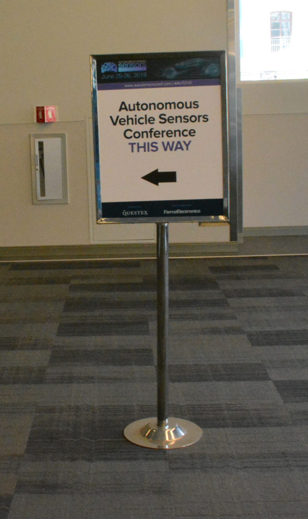 Hall sign on stanchion advertises, "Autonomous Vehicle Sensors Conference This Way"